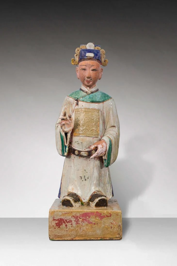 Tượng thị giả (người hầu) trong tư thế đứng trên bệ hình vuông, đầu đội mũ phốc vuông đắp nổi viên ngọc và mây. Mặt để mộc, dùng sơn để vẽ chân mày và mắt, tai to, trên mặt tạo 5 lỗ để gắn râu. Thân mặc áo dài màu trắng ngà, phần vai và ống tay áo màu xanh viền nâu, có đai, trên thân áo có “bổ tử” trang trí hình chim phượng, thủy ba, mây đối xứng trước và sau lưng. Tay trái cầm đai, tay phải đưa lên cao trước ngực trong tư thế cầm bút lông, chân mang hia.